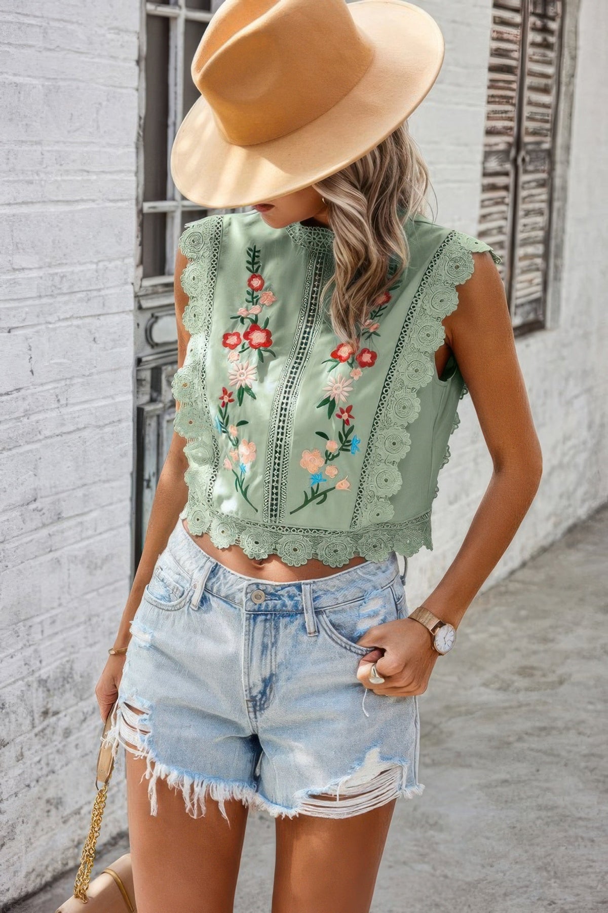 woman walking down street wearing green crochet top, cut off jean shorts and tan fedora hat.