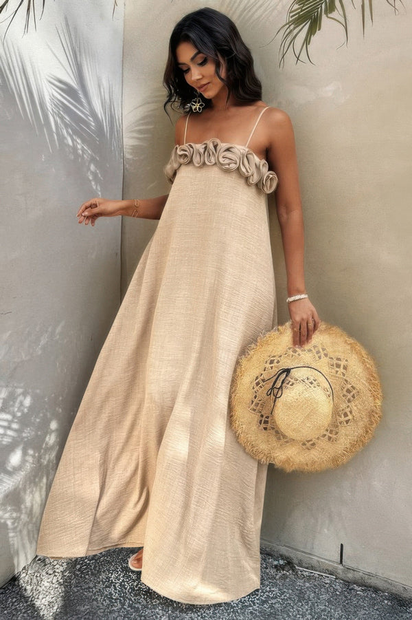 woman standing up holding sun hat, wearing beige color maxi dress with flower detail and spaghetti straps