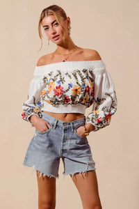 Woman wearing a floral off-shoulder top and denim shorts on a beige background