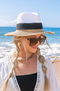 Woman wearing a white and black straw hat with sunglasses on a beach.