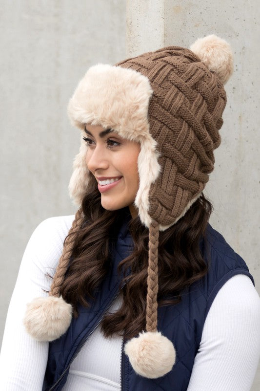 Woman wearing a brown knitted hat with fur trim and pom-poms against a gray background