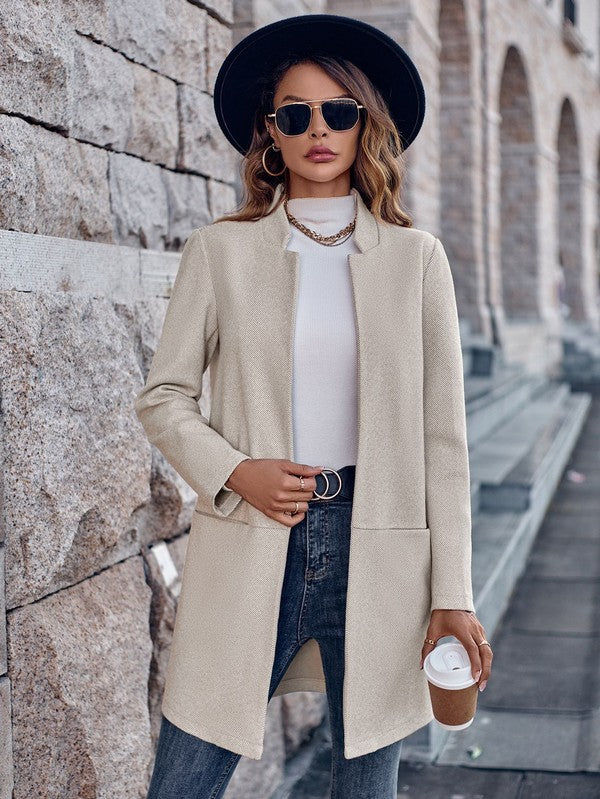 Woman in a beige coat and black hat standing against a stone wall.