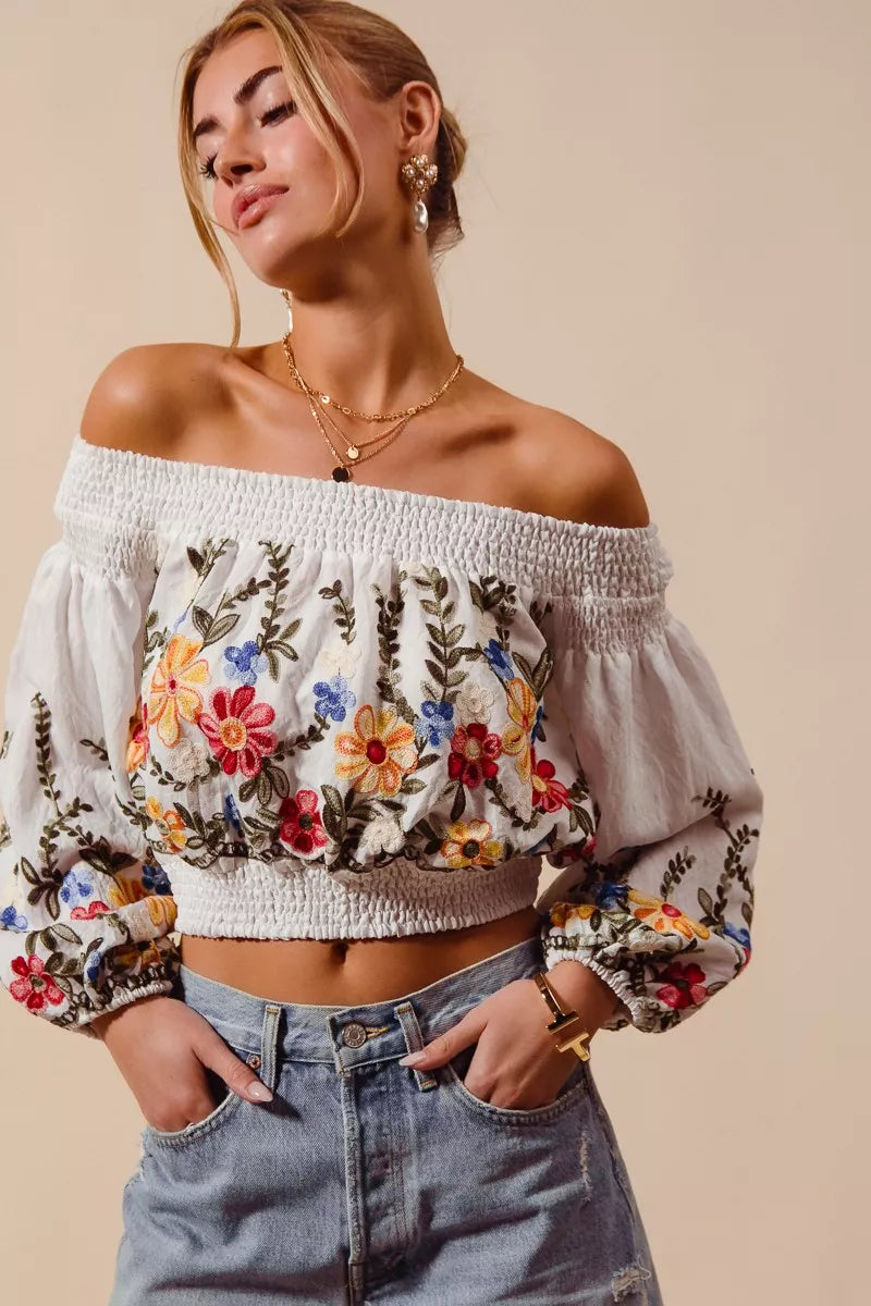 Woman wearing a floral embroidered off-shoulder top and jeans on a beige background