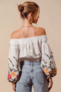 Woman wearing an off-shoulder blouse with floral embroidery and light blue jeans on a beige background