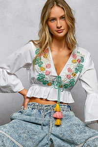 White cotton boho floral embroidered cropped V-neck blouse with braided tie, tassels, and flared ruffle sleeves.