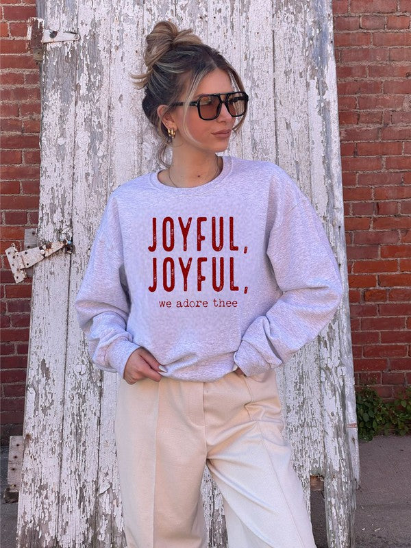 Person wearing a light gray sweatshirt with 'JOYFUL, JOYFUL, we adore thee' text against a rustic wooden and brick wall.