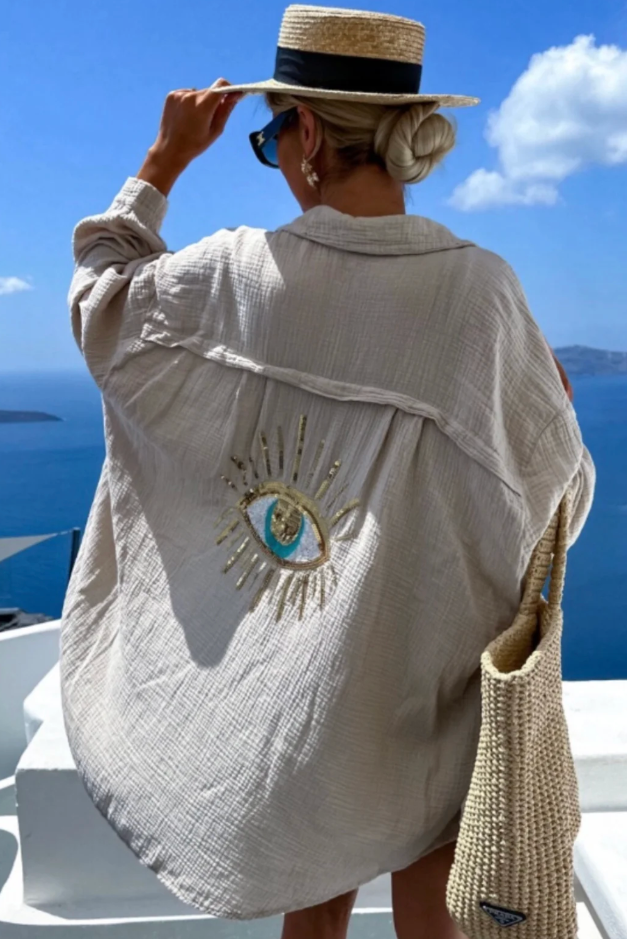 Person wearing a beige jacket with an eye design, standing against a blue sky and ocean backdrop.